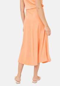 Jupe midi fluide couleur orange clair, fabriquée en tissu doux. Possède une coupe évasée flatteuse et une texture lisse, adaptée à un usage casual.