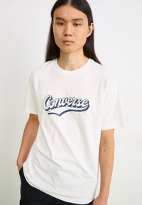 Hvit bomulls T-skjorte med et stort blått "Converse"-logo i kursiv skrifttype. Korte ermer og rund hals.
