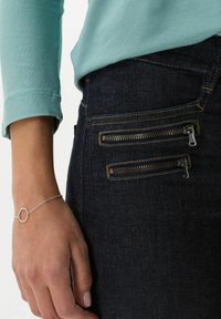 Jeans de mezclilla negros con bolsillos con cremallera y costuras doradas, combinados con una blusa de manga larga en verde menta. Una delicada pulsera de plata adorna la muñeca.