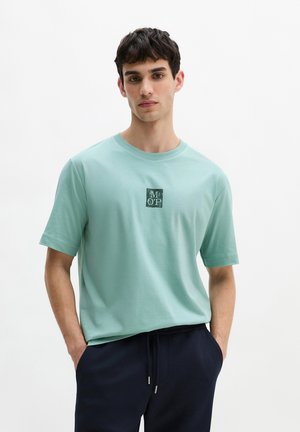 Giovane uomo con maglietta azzurra chiara con logo "M O P" e pantaloni scuri, in piedi con le mani in tasca su sfondo uniforme.