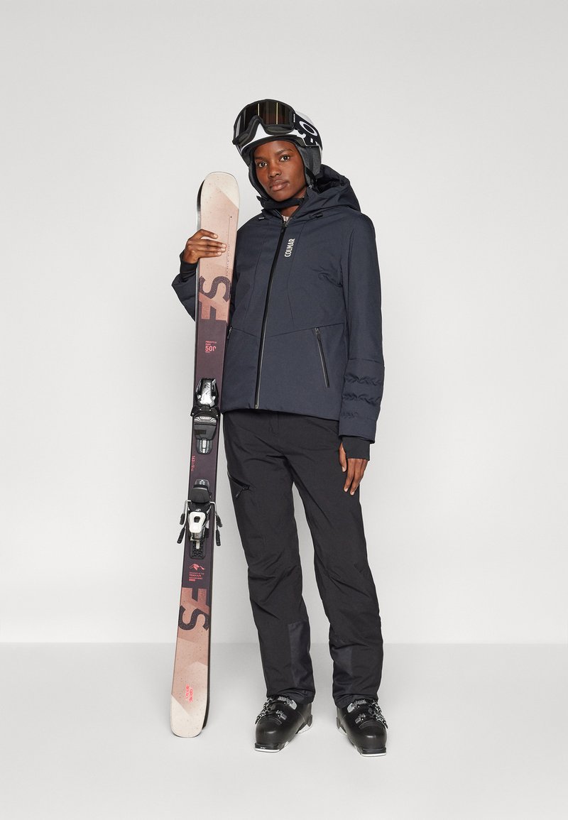 Die Skiausrüstung umfasst eine schwarze isolierte Jacke mit Reißverschlusstaschen, schwarze Hose und Skier mit einem braunen und rosa Farbverlauf, die schwarze Bindungen haben.