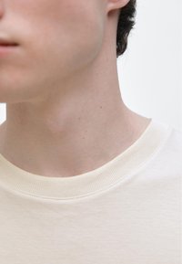 Beige bomullst-shirt med ribbad rund halsringning, med en mjuk textur och minimala sömnadsdetaljer synliga längs kragen.