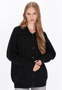 Svart stickad kardigan med krage, knappstängning framtill, ribbade muddar och nederkant, samt texturerade mönster på ärmarna.
