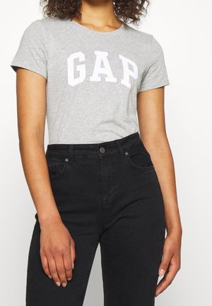 Kobieta w szarej koszulce z krótkim rękawem z logo "GAP" i czarnych, wysokich dżinsach na jednolitym tle.