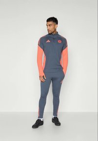 Sivi atletski trenirka s koraljnim naglascima. Dukserica s dugim rukavima i patentnim zatvaračem te uske hlače. Sadrži Adidas logo i pruge na ramenima i nogama.