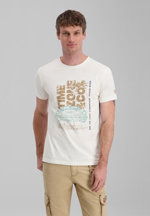 Mann trägt weißes T-Shirt mit Vintage-Auto und Schriftzug sowie beige Cargo-Hose, steht vor einfarbigem grauem Hintergrund.