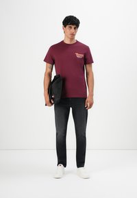 Junger Mann trägt ein burgunderfarbenes Tommy Jeans T-Shirt, schwarze Jeans, weiße Turnschuhe und hält eine schwarze Tasche vor einem schlichten weißen Hintergrund.