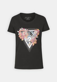 T-shirt en coton noir avec un graphique triangulaire comportant des motifs floraux et des pois blancs, mettant en avant le logo "GUESS" en lettres majuscules.