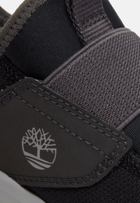 Nahaufnahme eines schwarz-grauen Sneakers mit strukturiertem Stoff, genähtem Lederteil und einem weißen Baumlogo an der Seite.