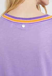 Blond persoon die een lavendelkleurig shirt draagt met een geribde halslijn, voorzien van paarse, oranje en roze strepen en een klein rond zilveren label aan de achterkant van de kraag.