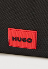 Material texturat negru cu o etichetă dreptunghiulară roșie, având în centru litere negre în relief "HUGO".