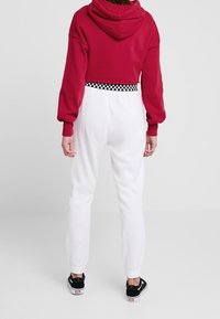 Felpa rossa corta con cappuccio, abbinata a pantaloni jogging bianchi con una vita a scacchi. Il modello indossa scarpe slip-on nere.