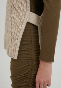 Beige gestrickte Weste mit einem strukturierten Muster, die eine seitliche Riemenbefestigung über einem enganliegenden olivgrünen Kleid mit gerafften Stoffdetails aufweist.