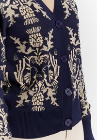 Cardigan lavorato a maglia con una base blu navy scuro e motivi floreali color crema, design a scollo a V e bottoni viola lungo il fronte.