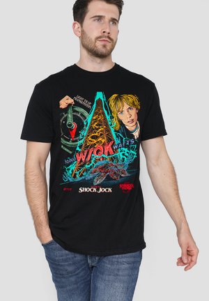 Mann trägt ein schwarzes Stranger Things Grafik-T-Shirt mit einem Mädchen, einem Strommast und einer Kreatur sowie dem Text "Shock Jock" und "WQK Watts".