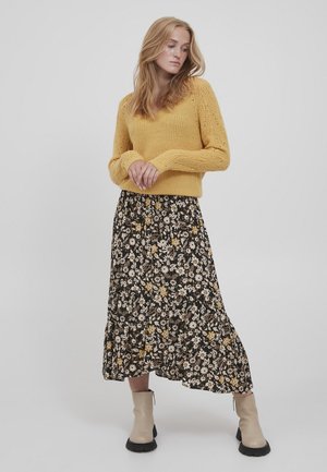 Gelber gestrickter Pullover mit V-Ausschnitt, kombiniert mit einem schwarzen Maxirock mit floralen Mustern in Weiß und Gelb. Beigefarbene Knöchelstiefel mit dicker Sohle.