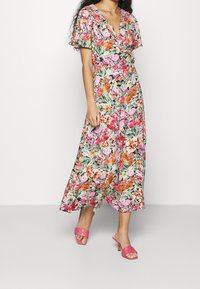 Femme portant une robe midi colorée à fleurs avec des manches courtes et des sandales à talons roses, se tenant devant un fond blanc uni.
