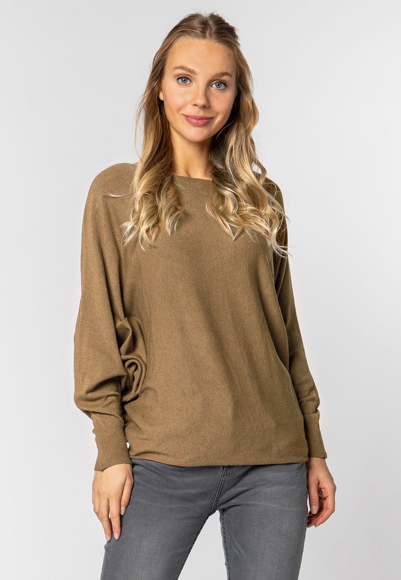 Bruine, losse trui met een ronde hals en lange mouwen, gecombineerd met grijze jeans, op een neutrale achtergrond. Soepele textuur.