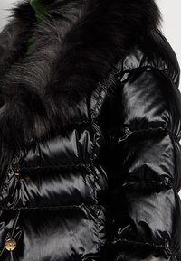 Chaqueta acolchada negra con textura brillante y un cuello de piel negra de peluche. Presenta herrajes dorados en el frente y un diseño acolchado horizontal.