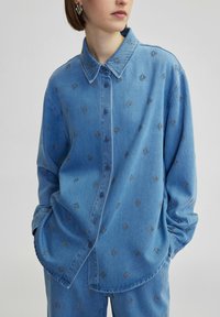 Camicia in denim azzurro chiaro con colletto a bottoni, caratterizzata da motivi a diamante ricamati e una vestibilità comoda. Maniche lunghe e tasche laterali.