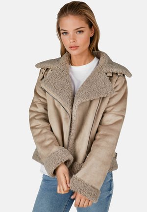 Junge Frau trägt eine beige Lammfelljacke mit großem Kragen und Manschetten, weißes Hemd und blaue Jeans, steht vor weißem Hintergrund.