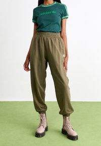 Žena oblečená v zeleném tričku Adidas, olivových teplácích Adidas a béžových šněrovacích kotníkových botách stojí na zelené podlaze.