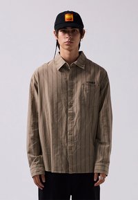 Long-sleeve, light brown striped shirt with a collar, front pocket, and button closure. Paired with a black cap featuring a logo patch.