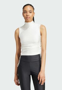 Witte mouwloze top met een hoge hals, ruches aan de zijkanten en trekkoorden, gecombineerd met zwarte gebreide leggings. Eenvoudig, modern ontwerp.