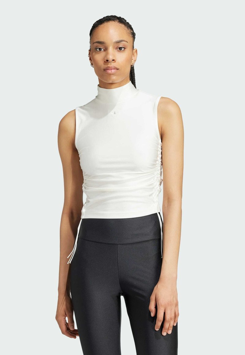Witte mouwloze top met een hoge hals, ruches aan de zijkanten en trekkoorden, gecombineerd met zwarte gebreide leggings. Eenvoudig, modern ontwerp.