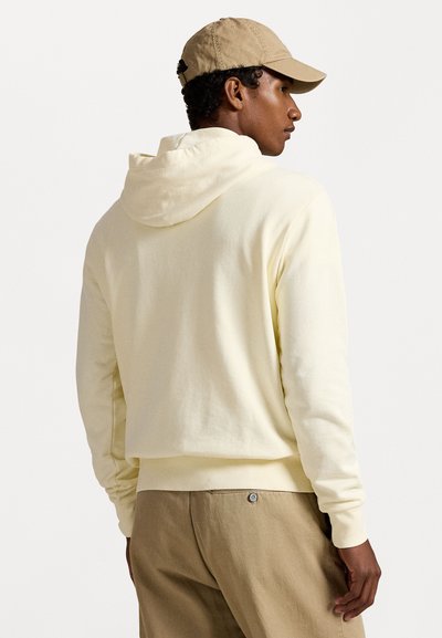 Gul bomullshoodie med hette og snøring, avslappet passform og ribbestrikkede mansjetter, kombinert med beige bukser. Enkelt design, uten mønstre eller logoer.