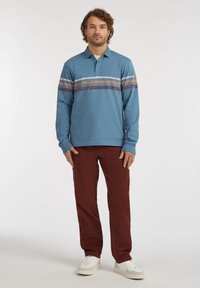 Blauwe polo met lange mouwen en horizontale meerkleurige strepen; gecombineerd met bordeauxrode broek en witte sneakers. Subtiele logo op de mouw.