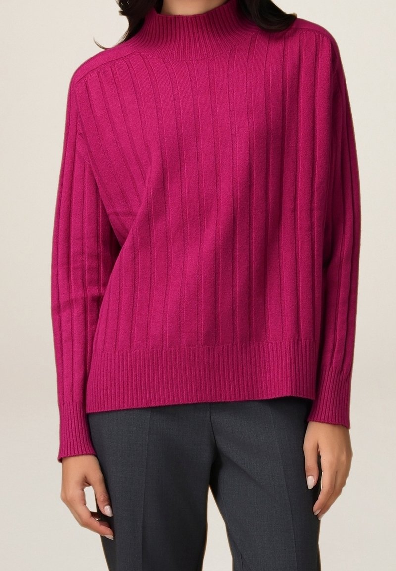 Pull côtelé fuchsia avec un col montant et une coupe à épaules tombantes, doté d'une texture douce et d'une coupe décontractée, associé à un pantalon gris.