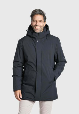 Homme souriant portant un manteau d'hiver noir à capuche, les mains dans les poches, debout devant un fond clair uni.