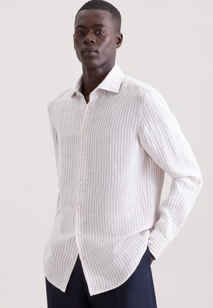 Homme portant une chemise à rayures verticales blanches et rose clair avec un pantalon foncé, posant avec une main dans la poche devant un fond uni.