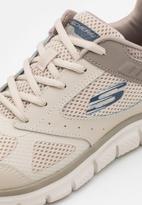 Zapatilla deportiva beige con paneles de malla, punta redondeada y suela texturizada. Logo prominente en el lateral, cordones en color a juego.