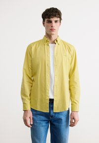 Νεαρός άνδρας με κοντά σγουρά μαλλιά, που φορά ανοιχτό κίτρινο πουκάμισο πάνω από λευκή μπλούζα και μπλε τζιν, στέκεται μπροστά σε απλό λευκό φόντο.