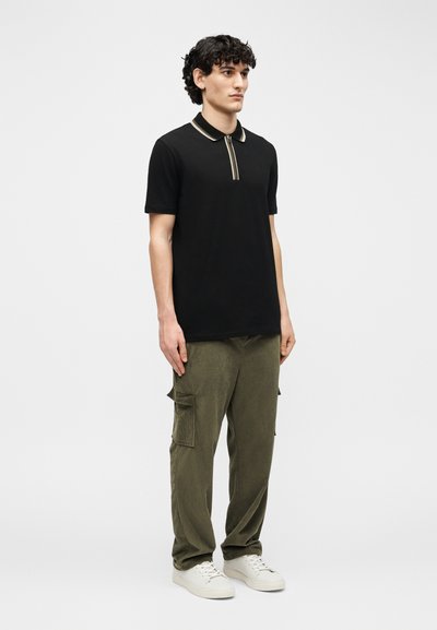 Νεαρός άνδρας που φοράει μαύρο πόλο με κοντά μανίκια, χακί παντελόνι cargo και λευκά αθλητικά παπούτσια, στέκεται μπροστά σε απλό φόντο.
