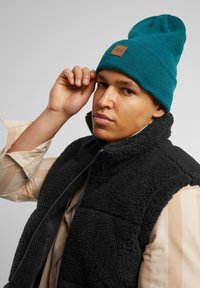 Beanie tricotat de culoare teal, cu o etichetă din piele maro, purtat cu o vestă din fleece neagră peste o cămașă cu dungi bej. Accent pe textură și pe aspectul stratificat.