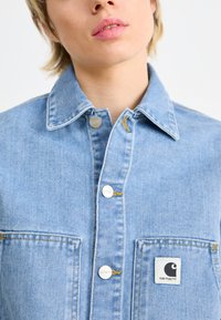 Hele sinine denim jakk, millel on klassikaline krae, hõbedased nööbid, kaks eesmist taskut ja silmatorkav Carhartti logo märk.