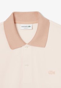 Polo paita pehmeässä vaaleanpunaisessa värissä, jossa on kontrastivärinen beige ribbineulekaulus, kaksinapitus ja brodeerattu logo vasemmassa rinnassa. Teksturoitu kangas.