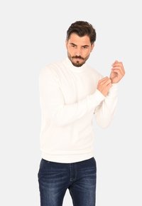 Maglione bianco a collo alto in morbido materiale lavorato a maglia con polsini e orlo a coste, abbinato a jeans denim blu scuro. La modella sta aggiustando le maniche.
