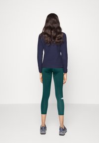 Granatowa bluzka z długim rękawem w połączeniu z zielonymi legginsami sportowymi z logo. Model nosi szare sneakersy, widok od tyłu.