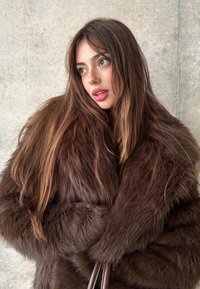 Brun pälsrock med glansig struktur, med vida ärmar och voluminös design. Långt, vågigt hår som delvis täcker ansiktet.