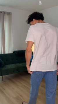 Tânăr cu păr creț stă în sufragerie cu fața spre departe, purtând un tricou alb și blugi albaștri lângă o canapea verde închis și draperii.