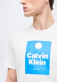 Νεαρός άνδρας που φοράει λευκό μπλουζάκι με μπλε τετράγωνο λογότυπο και λευκό κείμενο «Calvin Klein», στραμμένος προς τα αριστερά σε απλό φόντο.