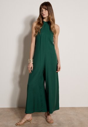 Donna in piedi che indossa una tuta a gamba larga verde scuro senza maniche con collo allacciato al collo e sandali beige con tacco e cinturini, braccialetti beige al polso.