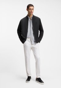 Άνδρας που φορά μαύρο μπουφάν bomber πάνω από ανοιχτό μπλε πουκάμισο, λευκό παντελόνι και μαύρα αθλητικά παπούτσια, στέκεται με τα χέρια στις τσέπες.