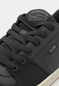 Black sneaker featuring a mix of smooth and textured materials, round laces, perforated accents, and a white rubber sole. Brand logo on tongue.