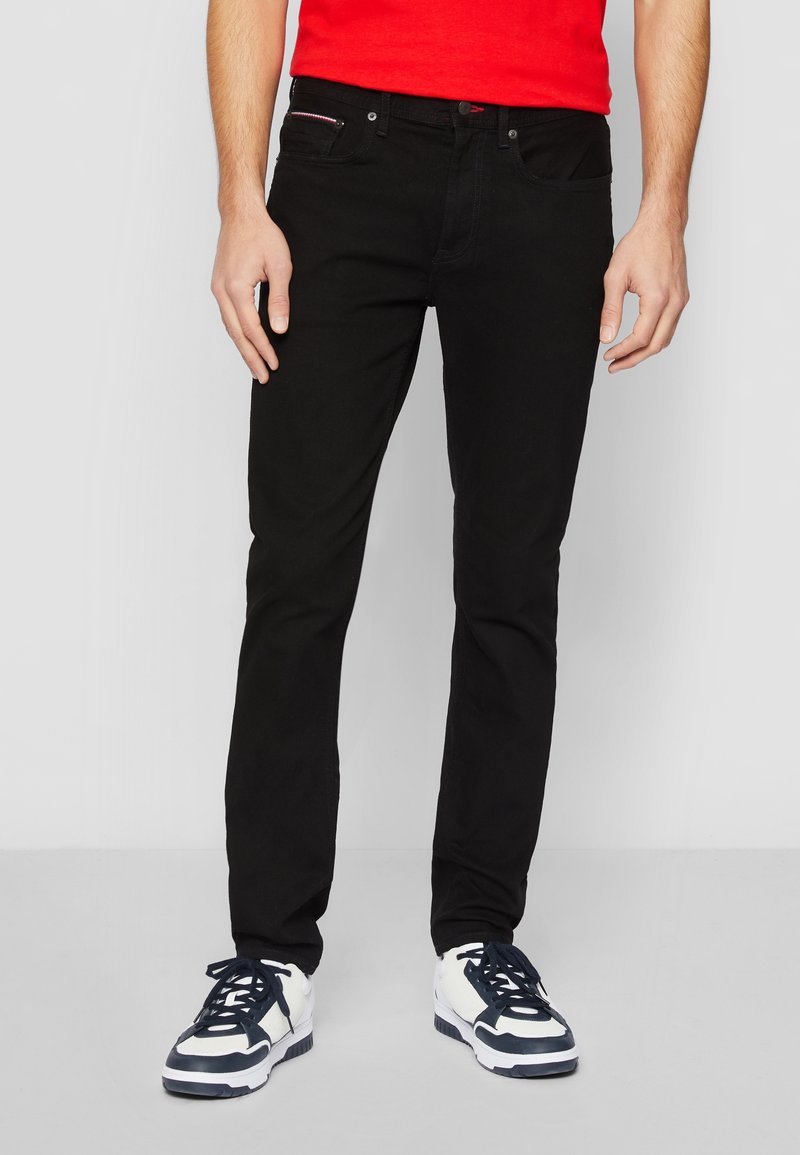 Hombre vistiendo pantalones vaqueros slim-fit negros, zapatillas blancas y negras, y una camisa roja, de pie contra un fondo liso de color gris claro.