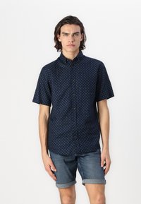 Chemise bleu marine à manches courtes avec des pois blancs, col boutonné et coupe décontractée. Associée à un short en denim retroussé.